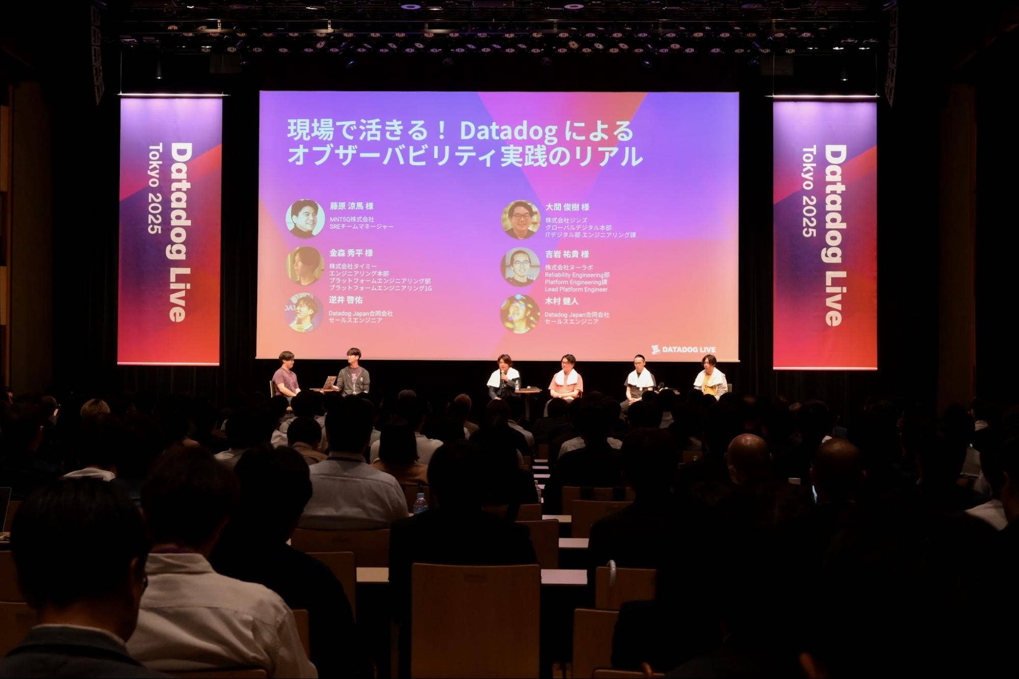 セッションタイトルと登壇者名を表示したスクリーンの前で、観客に向かってステージに座る2人のモデレーターと4人のパネリスト。